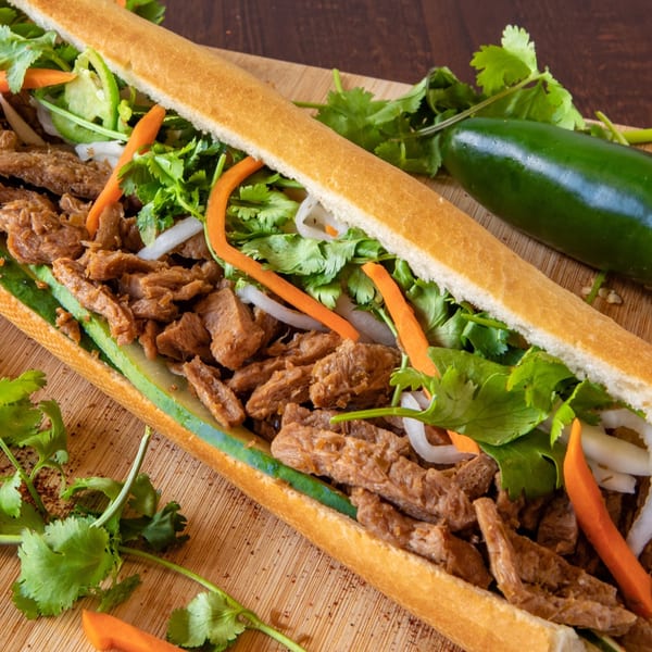 A Paris Banh Mi sandwich with meat, cilantro, carrots, and cucumber on a wooden board.