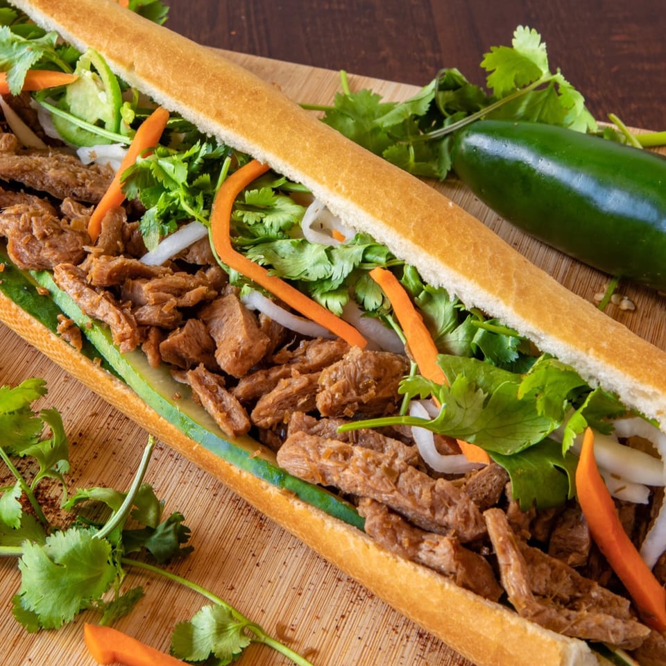 A Paris Banh Mi sandwich with meat, cilantro, carrots, and cucumber on a wooden board.
