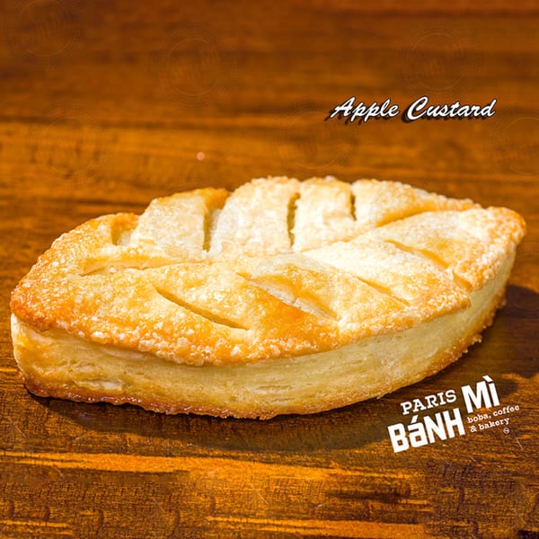 Golden-brown apple custard pastry with a leaf-like design, dusted with powdered sugar on a wooden surface.