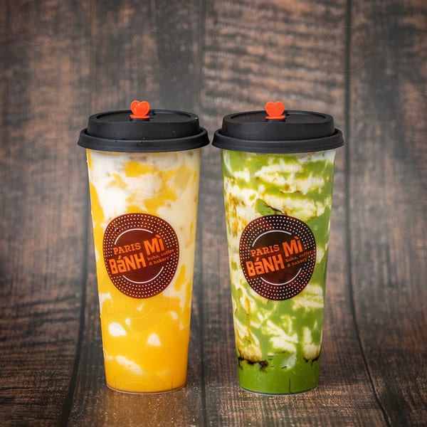 Two Paris Banh Mi boba drinks, one yellow and one green, on a wooden surface.