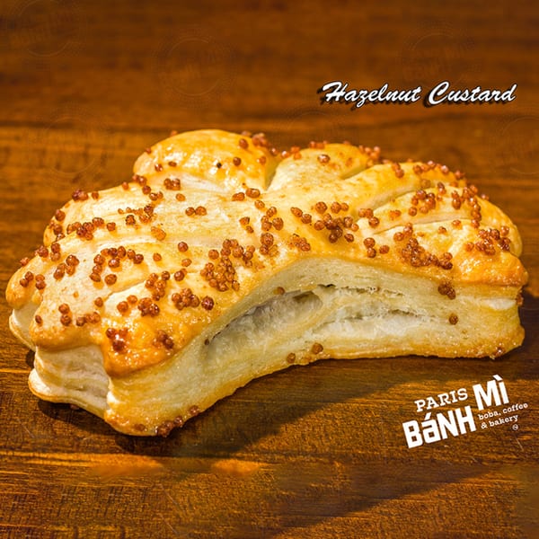 A golden-brown hazelnut custard pastry, sprinkled with crunchy toppings, rests on a wooden surface.
