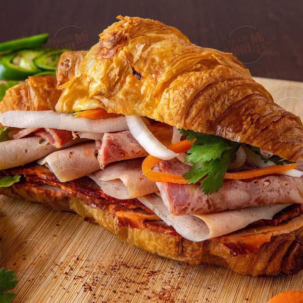 A Paris Banh Mi croissant sandwich filled with various meats, pickled vegetables, and cilantro on a wooden board.